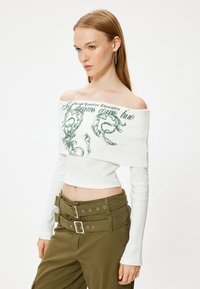Weißes Off-Shoulder-Top aus geripptem Stoff mit grüner Grafikschrift. Kombiniert mit hochgeschnittenen olivgrünen Hosen mit Doppelverschluss.