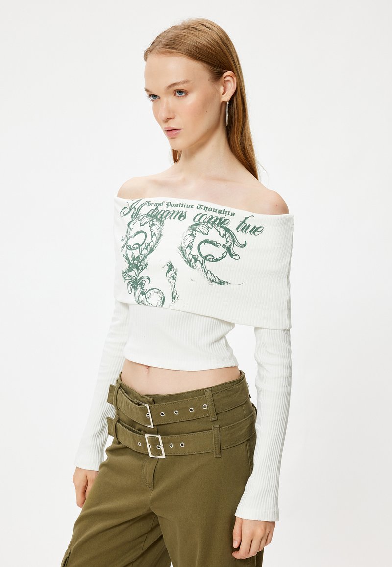 Weißes Off-Shoulder-Top aus geripptem Stoff mit grüner Grafikschrift. Kombiniert mit hochgeschnittenen olivgrünen Hosen mit Doppelverschluss.