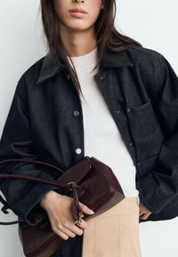 Dark denim jacket with a black collar, button front, and a chest pocket, paired with a fitted white ribbed top and a maroon handbag.