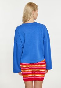 Kék, oversized pulóver, ejtett vállakkal, multicolor cikk-cakk mintázatú kötött szoknyával párosítva, élénk piros, rózsaszín, narancs és sárga csíkokkal.