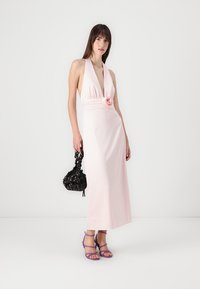 Maxiklänning i ljusrosa satin med halterneck, samlad midja och tygrosdetalj; bärs med en svart väska och lila remsandaler.