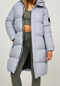 Hellgraue Steppjacke mit Kapuze, ausgestattet mit einem Frontreißverschluss, Seitentaschen und einem gesteppten Muster. Das Innenfutter ist schwarz mit einem leichten Glanz.