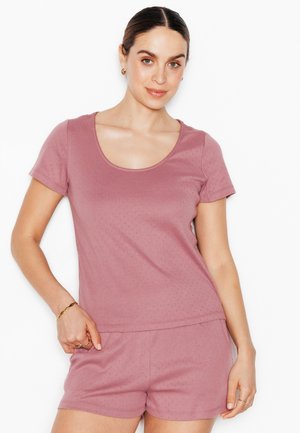 Donna con capelli scuri con un top rosa a maniche corte e pantaloncini abbinati, in piedi con una mano sui fianchi su sfondo bianco.