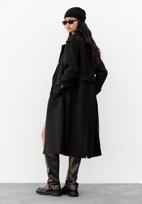 Crni vuneni kaput s remenom u struku, bočnim džepovima i slobodnim krojem. Uparen s crnim čizmama do koljena koje imaju metalne kopče.