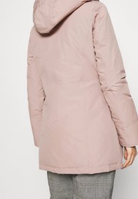 Woman wearing a light pink hooded winter coat and gray checkered pants, shown from the back against a plain background.