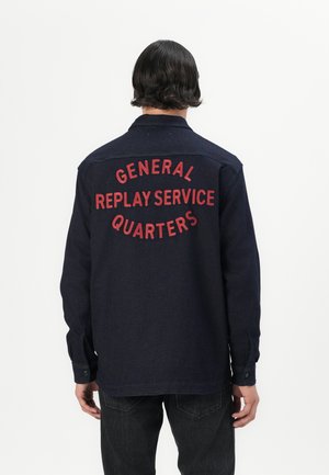 Camicia blu scuro con testo ricamato rosso che dice "SERVIZIO GENERALE REPLAY QUARTI" sulla schiena. Maniche lunghe e polsini con bottoni.