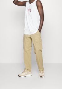 Vit ärmlös skjorta med röd text, beige cargobyxor med fickor och beige New Balance-sneakers. Neutral bakgrund.