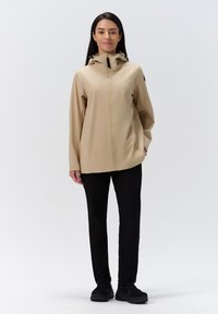 Beige buitenjacket met een ritssluiting aan de voorkant en een capuchon, gemaakt van glad materiaal. Gepaarde met zwarte broek en zwarte sportschoenen.