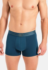 Pánské boxerky v tmavě modrozelené bavlně s elastickým pasem nesoucím logo CR7 a zlaté pruhy. Hladká textura s přiléhavým střihem.