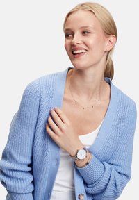 Hellblauer, lockerer Strickcardigan mit ribbelten Bündchen, über einem weißen Oberteil getragen. Silberne Uhr und zarte Halskette mit kleinen Perlen.