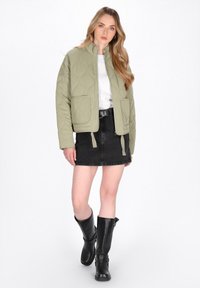 Quiltet olivengrønt jakke med høj krave, to forlommer, cropped design, parret med en sort denimnederdel og knæhøje sorte støvler.