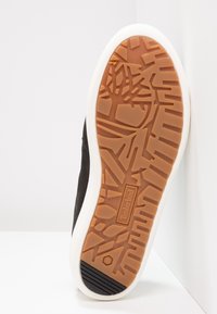 Semelle en caoutchouc d'une chaussure noire avec des motifs géométriques sur la semelle et le logo Timberland au centre. Une semelle intermédiaire blanche entoure la semelle.