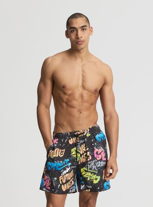 Giovane uomo con fisico tonico che indossa pantaloncini da bagno neri con scritte e disegni colorati in stile graffiti, in piedi davanti a uno sfondo semplice.