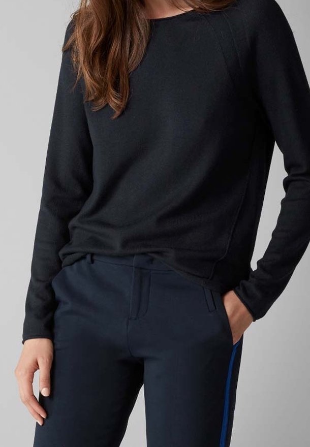 Femme portant un haut noir à manches longues et un pantalon bleu marine avec des bandes latérales bleues, main dans la poche, sur un fond gris uni.