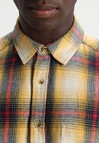 Camicia a scacchi gialla con accenti blu e rossi, dotata di colletto standard e due bottoni visibili al collo.
