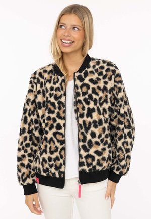 Junge Frau lächelt, trägt eine beige-schwarze, flauschige Jacke mit Leopardenmuster, schwarzen Bündchen und weiße Hose vor weißem Hintergrund.