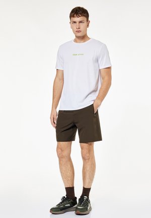 Młody mężczyzna stoi z rękami w kieszeniach, ubrany w białą koszulkę z napisem „KEEP ACTIVE”, ciemne krótkie spodenki, brązowe skarpety i zielone buty sportowe na białym tle.