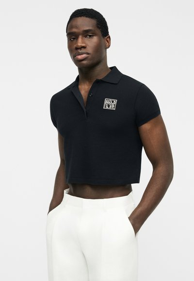 Homme portant un polo noir court avec un logo sur la poitrine et un pantalon blanc taille haute, les mains dans les poches, sur fond uni.