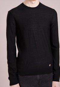 Svart långärmad tröja i ett mjukt, texturerat material. Har en rund hals och en liten metallisk logga i det nedre vänstra hörnet.