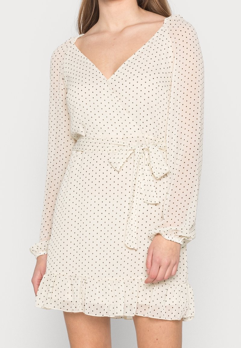 Robe portefeuille en tissu crème transparent, présentant des pois noirs, des manches longues, une taille nouée et un ourlet froncé. Légère et texturée.