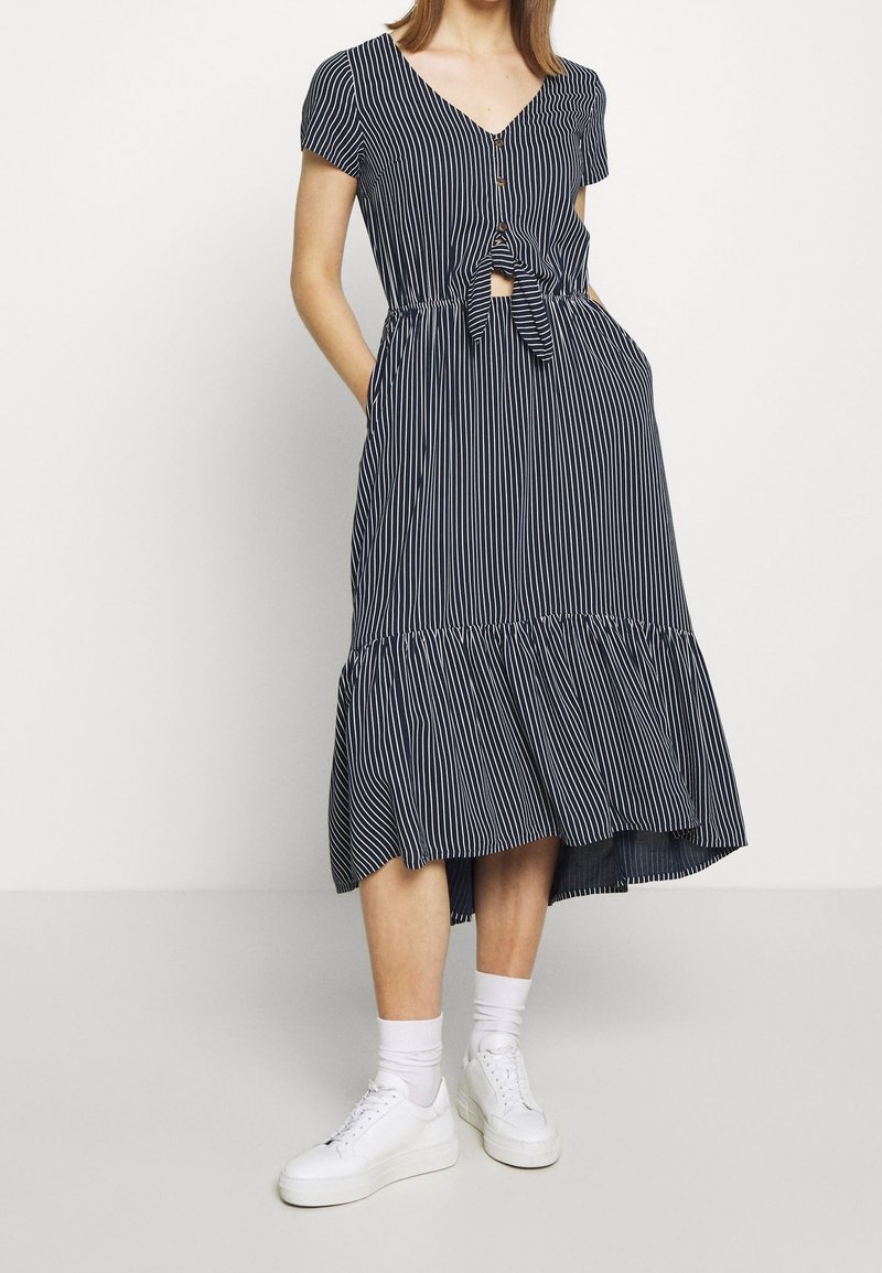 Abito a righe blu navy con maniche corte, scollo a V, dettaglio dei bottoni, cintura in vita e orlo arricciato. Abbinato a sneakers bianche e calzini alla caviglia.