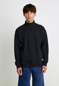 Sudadera negra con cremallera y alto cuello, con un discreto logo bordado en el pecho, combinada con pantalones de denim azul holgados.