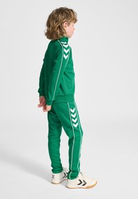 Copil cu păr creţ stă în profil, purtând un trening verde cu dungi albe şi pantofi sport albi, pe un fundal simplu.