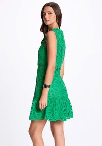 Robe en dentelle verte avec un corsage ajusté, des bords festonnés et des motifs découpés floraux. Design sans manches avec une jupe évasée.