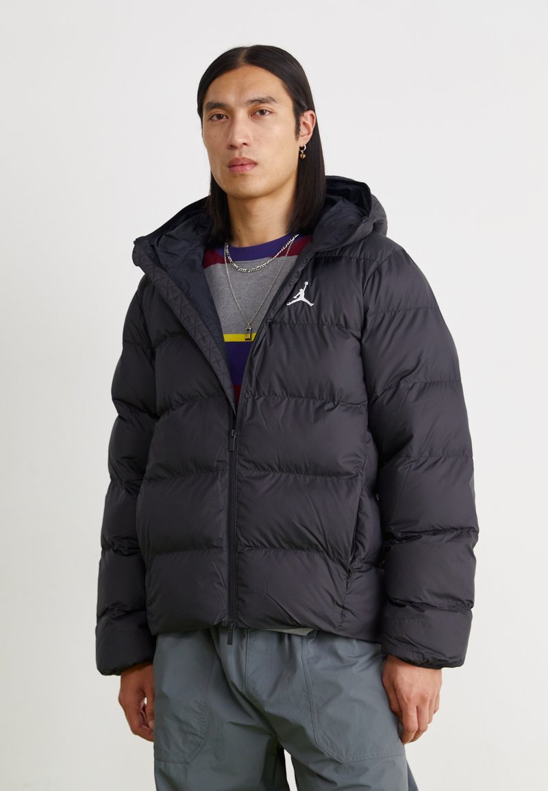 Black puffer jacket with a hood, featuring quilted sections and a front zipper. White logo on the left chest, worn over a gray striped shirt.