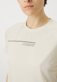 Lys beige kortermet T-skjorte med ribbestruktur og en mørkegrå "HUMMEL" logo sentrert under et horisontalt linjedetalj.