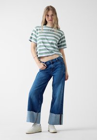 T-shirt cropped a righe verdi e bianche abbinato a jeans a gamba larga blu con orli sfrangiati e sneakers bianche. Design semplice.