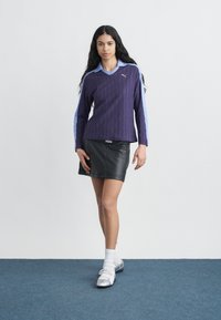 Γυναίκα με μακριά σκούρα μαλλιά που φοράει μοβ πουλόβερ Puma, μαύρη δερμάτινη φούστα, λευκές κάλτσες και ασημένια παπούτσια, που στέκεται σε μπλε χαλί.