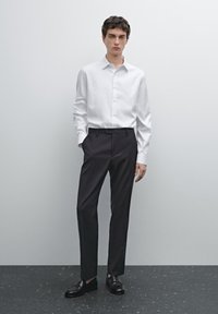 Weißes Langarm-Hemd mit Knöpfen, maßgeschneiderte dunkle Hosen und schwarze Slipper, stehend auf einer strukturierten dunklen Oberfläche vor einem hellen Hintergrund.