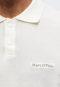 Κοντινό πλάνο ενός ατόμου που φοράει λευκό πόλο με δύο κουμπιά και κεντημένο το "Marc O'Polo" στο στήθος.