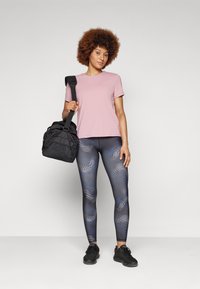 Pink short-sleeve shirt, patterned black and blue leggings, and black sneakers. Model holds a black gym bag, standing against a gray background.