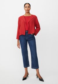 Rote, strukturierte, kurz geschnittene Jacke mit Reverskragen, kombiniert mit einem taillierten roten Oberteil und dunkelblauen, weit geschnittenen Jeans. Schwarze, hochhackige Pumps.