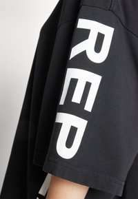 Černé tričko vyrobené z bavlny, které má na rukávu výrazný bílý text. Design zdůrazňuje uvolněný střih s krátkými, mírně oversized rukávy.