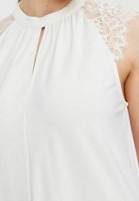 Blusa bianca con colletto alto, dettagli plissettati e accenti in pizzo sulle spalle. Tessuto leggero con una texture morbida.
