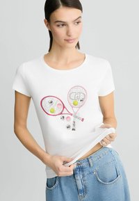 T-shirt blanc à manches courtes avec des raquettes de tennis imprimées, des balles de tennis colorées et des détails décoratifs, associé à un short en denim bleu.