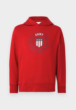 Red hoodie made of cotton with a logo featuring a shield, stars, and stripes in navy and white. Features a drawstring hood and ribbed cuffs.