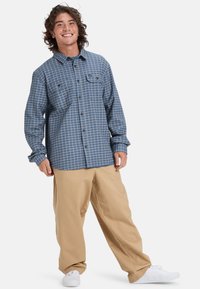 Blauw geruit overhemd met knopen en een borstzak, gecombineerd met losse beige broeken en witte sneakers. Het materiaal lijkt casual en gestructureerd.
