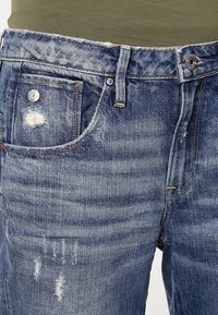 Jean en denim bleu avec de petites imperfections, poches avant visibles, passants de ceinture et fermeture à bouton métallique.