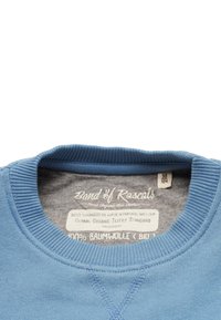 Blå bomullströja med ribbad krage, med en grå innerväv. Etiketterna anger ekologiskt material och produktionsstandarder.