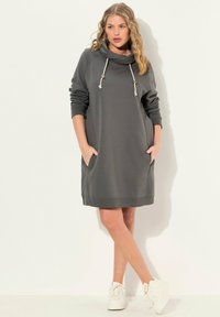 Graues Kapuzenkleid mit einer Schalkragen, langen Ärmeln, seitlichen Taschen sowie gerippten Bündchen und Saum. Kombiniert mit weißen Sneakers.