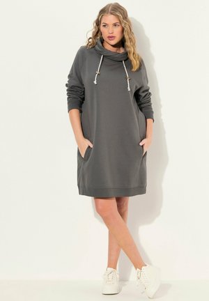 Robe sweat à capuche grise avec un col drapé, des manches longues, des poches latérales et des bords côtelés. Associée à des baskets blanches.