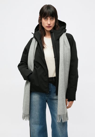 Crna izolirana jakna s kapuljačom, uparena sa svijetlosivim šalom. Ima prednji patentni zatvarač i opuštene plave traperice širokih nogavica.