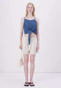 Haut cropped sans manches bleu avec un détail de nœud à l'avant, associé à un short rayé beige clair. Le mannequin porte un sac à main blanc et des sandales noires.