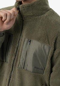 Fleec jakna u maslinastoj boji s visokim ovratnikom. Ima dva džepa sa zatvaračem, jedan sa teksturiranim materijalom, a drugi s glatkim, s brandom "PEGADOR."