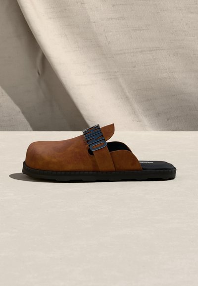 Mule en cuir brun à enfiler avec semelle en caoutchouc noire et bride ajustable ornée de lettres métalliques noires formant le mot "HOME".