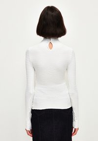 Maglietta bianca a maniche lunghe con tessuto a trama. Presenta un colletto alto e un piccolo foro a forma di chiave sulla schiena. Abbinata a una gonna in denim scuro.